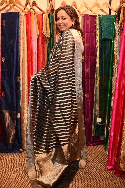 A woman standing and smiling, draped in a black and silver stripe zari silk sari, with a variety of colorful saris displayed in the background.